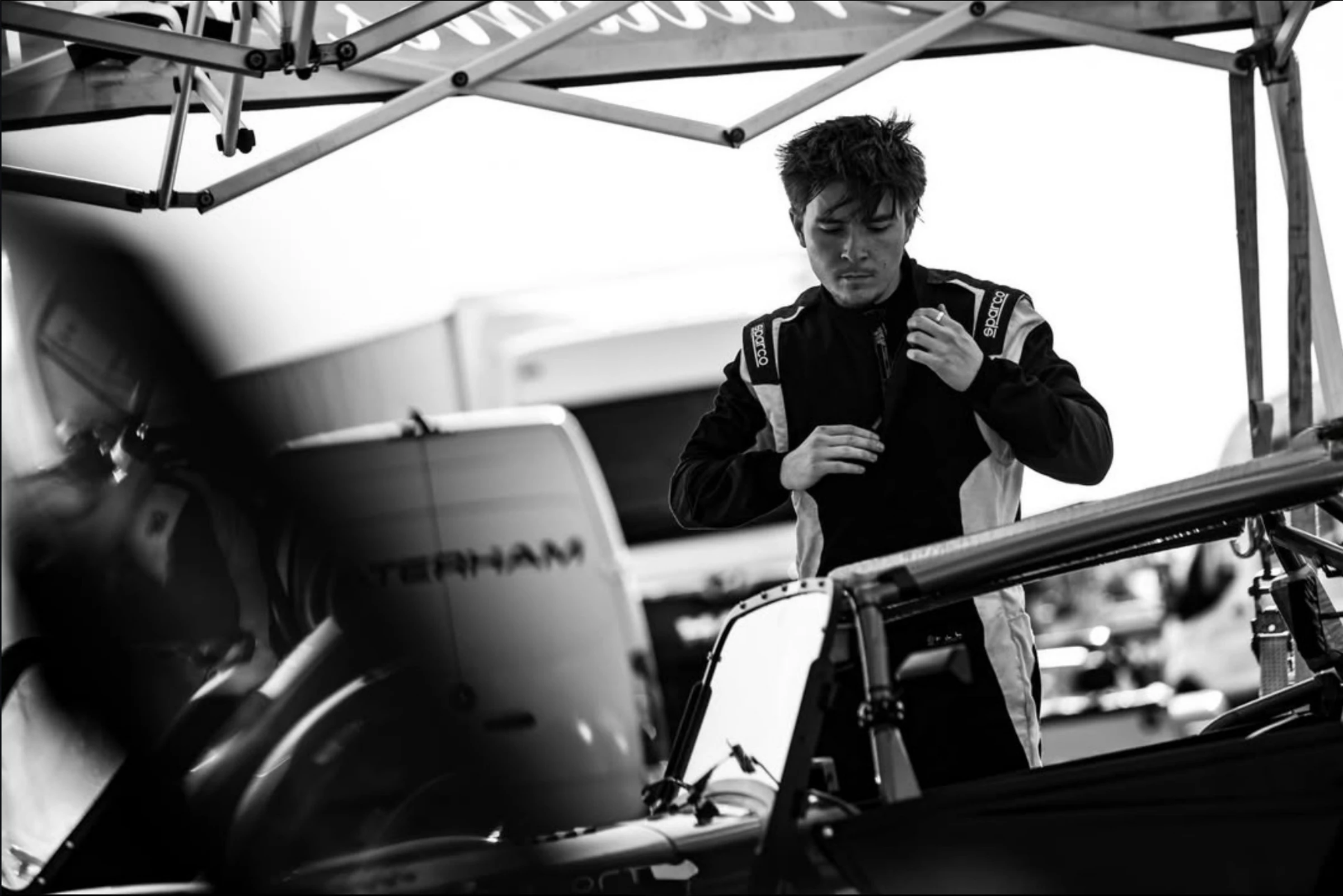 The driver pulling on his race suit in the paddock beside the car, in black and white.
